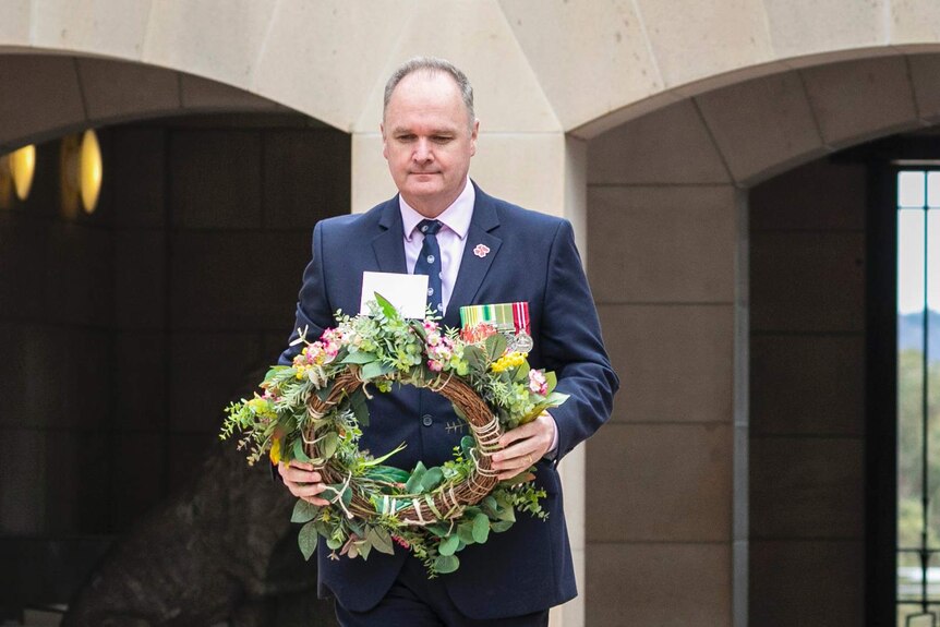 Man in a navy suit with a flower wreath walking forward with a stone archway in the background.