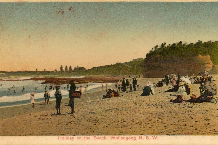 Postcard from the early 1900s showing people on a beach full dressed in heavy garments and shoes and ladies with parasols