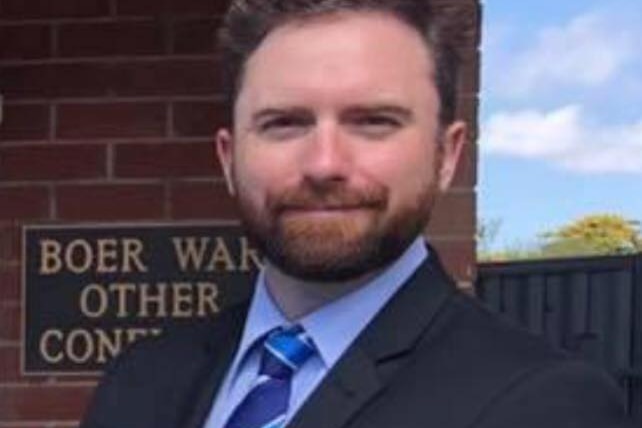 A closeup of a man with a short beard who is standing in front of a brick wall