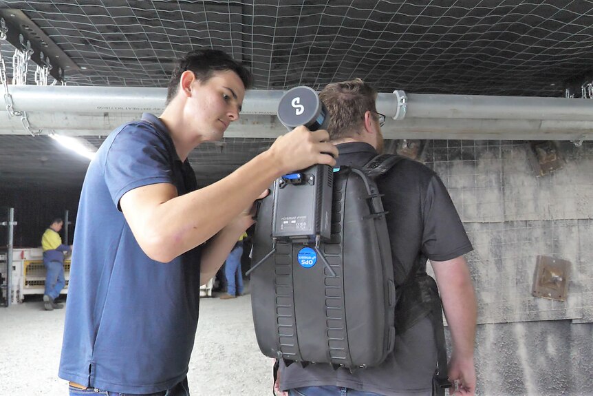 A man attaching a scanning device to a specialised backpack, being worn by another man.