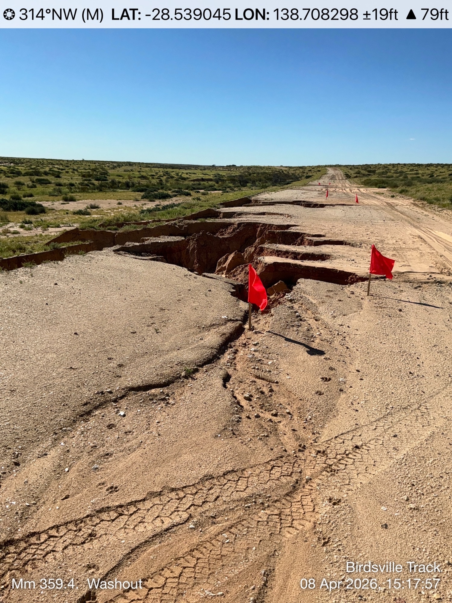A dirt road with a large chunk missing and flags marking the hole.