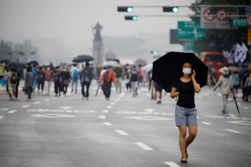 A young woman in black shirt and shorts walks along street with umbrella and mask with march behind.