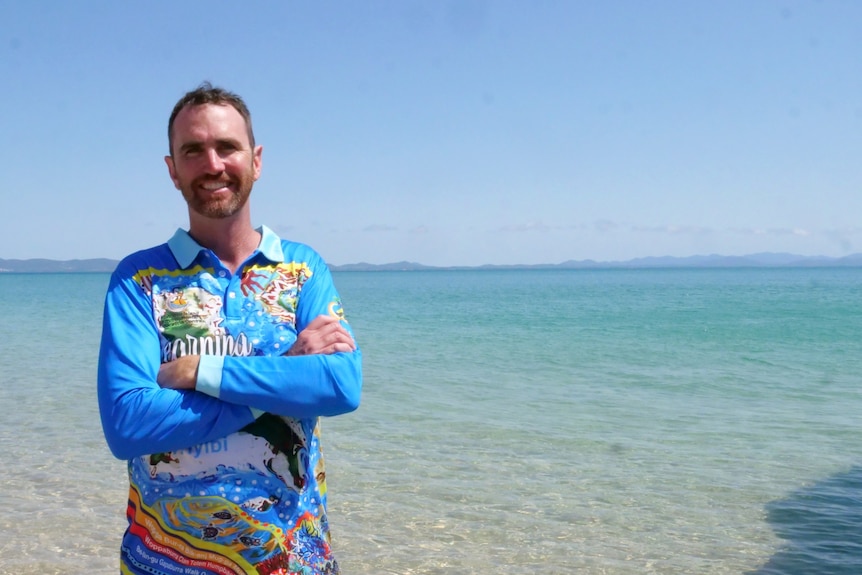A man stands in clear blue water, wearing a long sleeve blue shirt with his arms crossed and smiles.