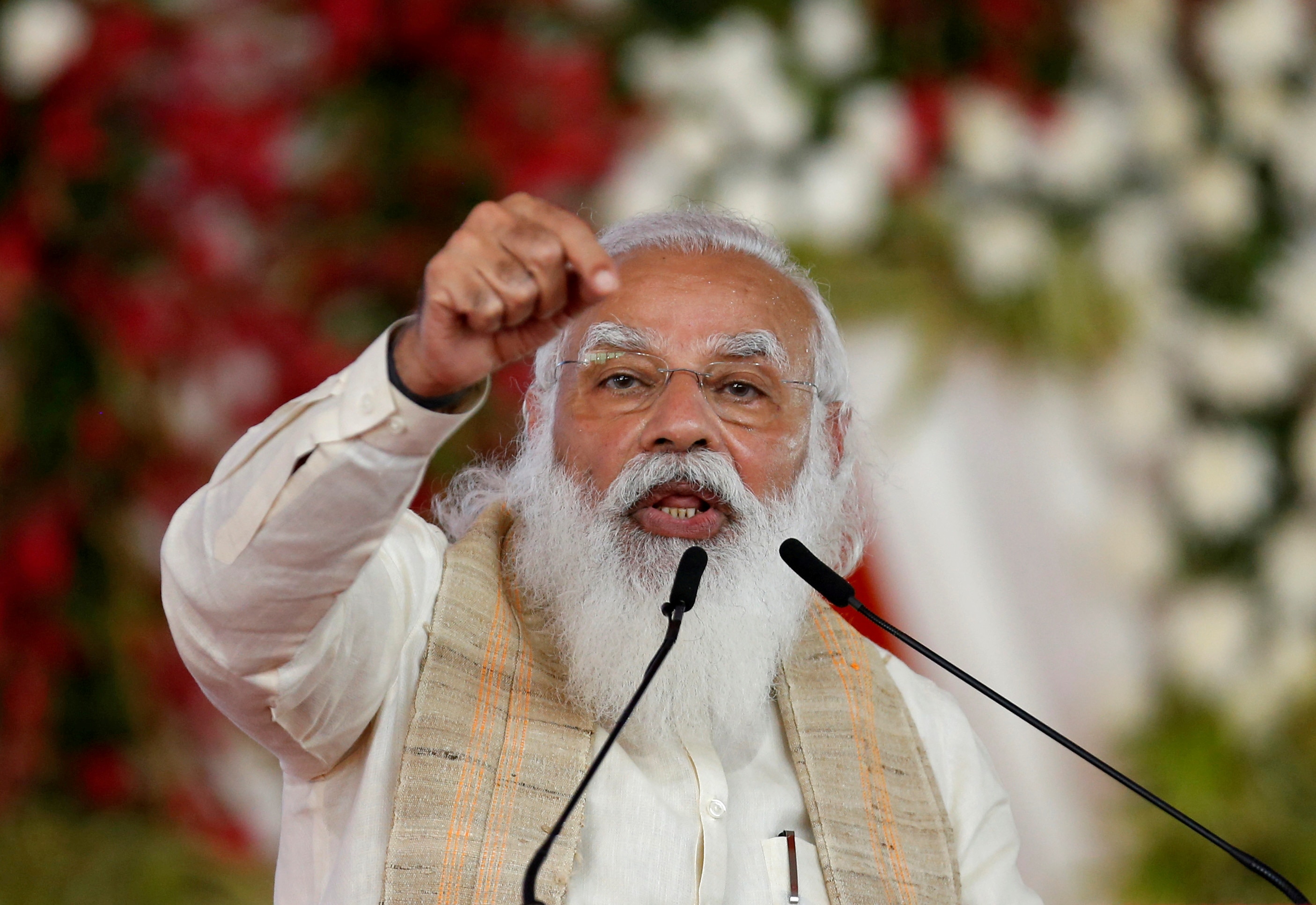An Indian man with a white beard speaks at a podium with his pointed hand in the air. 