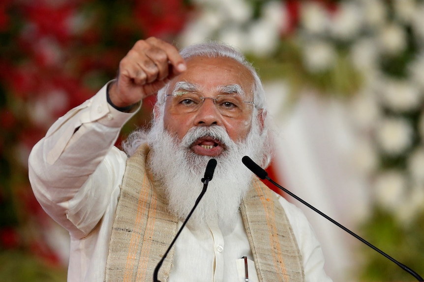 An Indian man with a white beard speaks at a podium with his pointed hand in the air. 