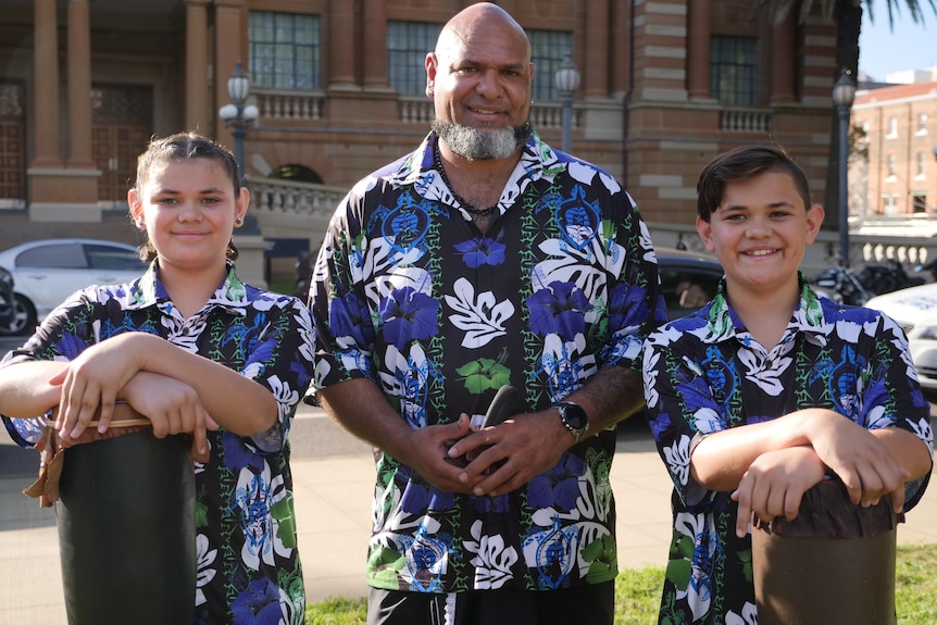 Torres Strait Islander man wearing blue, black, green and white shirt with two young boys standing either side.