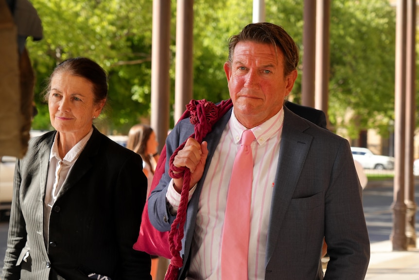 A man in grey suit and pink tie and a woman in a black suit and white shirt walk past court building.
