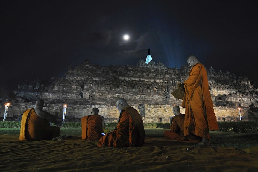 Five Buddhist monks in orange robes pray in front of the ancient Borobudur temple with a full moon.