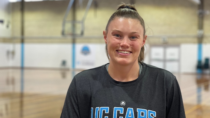 A woman with brown hair in a pony tail smiles. She's wearing a black Canberra Capitals T-shirt and is standing in a gymnasium