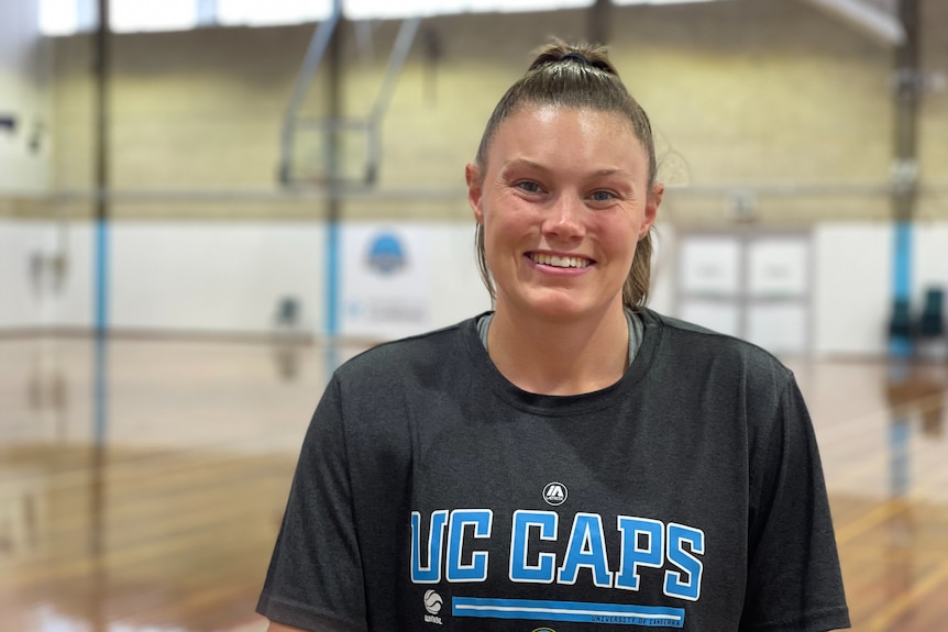 A woman with brown hair in a pony tail smiles. She's wearing a black Canberra Capitals T-shirt and is standing in a gymnasium