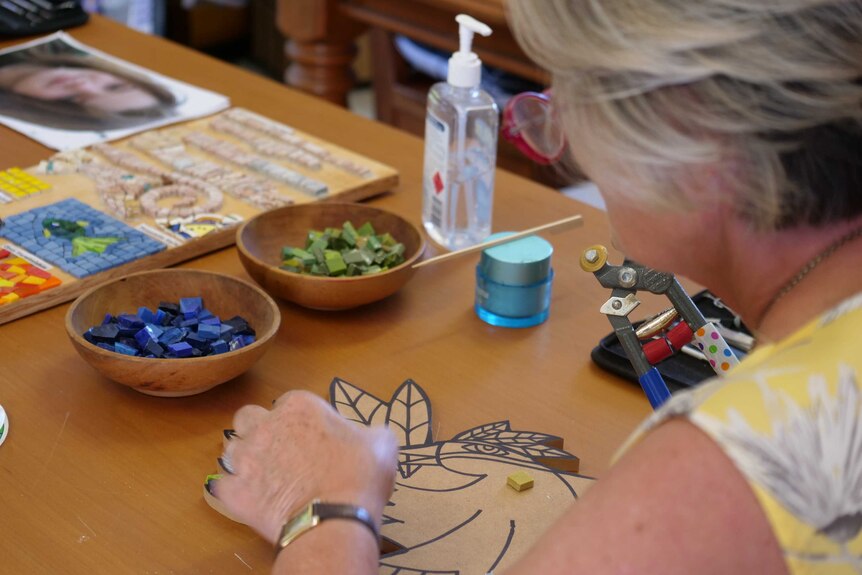 A woman uses coloured tiles to create a mosaic on a wooden table.
