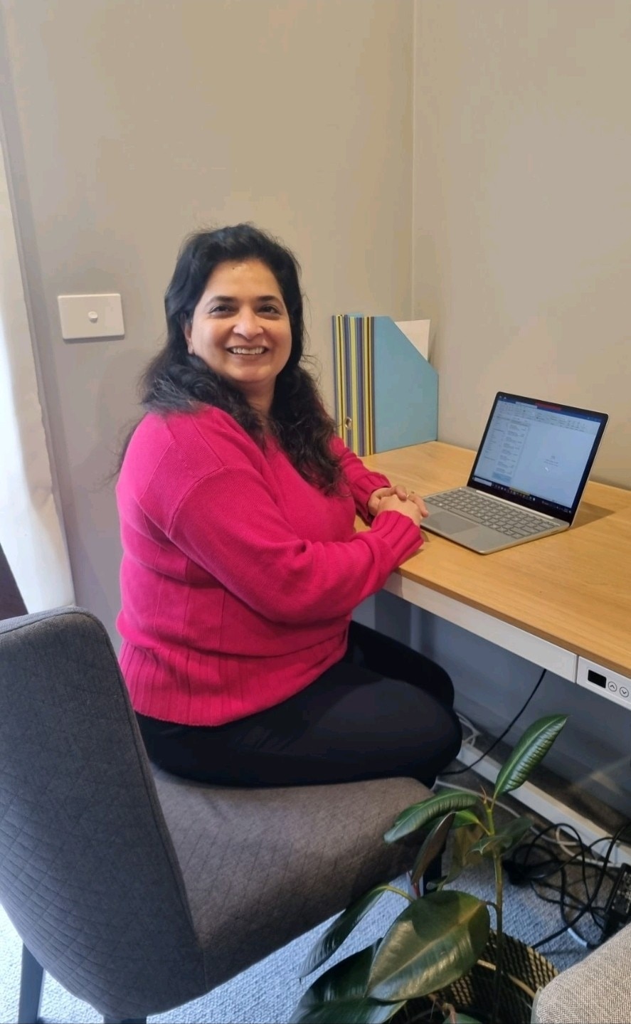 A woman with black hair and in a pink jumper, sits at her laptop on a desk, smiling at the camera. 