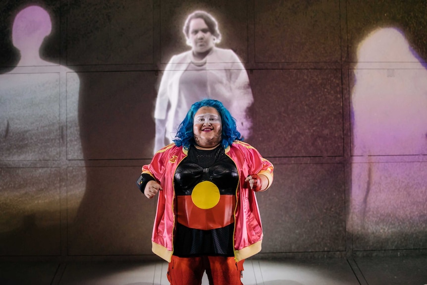 The actor stands, wearing a blue wig, facial makeup and a bustier emblazoned with the Aboriginal flag.