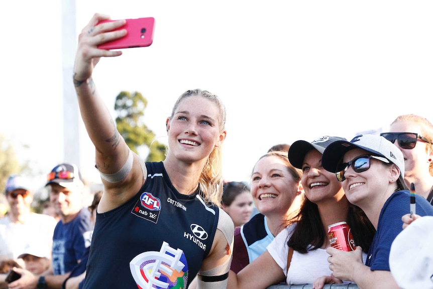 Tayla Harris smiles and holds up a phone to take a selfie with fans on the sidelines