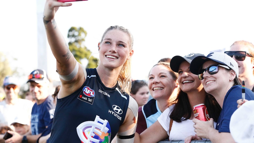 Tayla Harris smiles and holds up a phone to take a selfie with fans on the sidelines