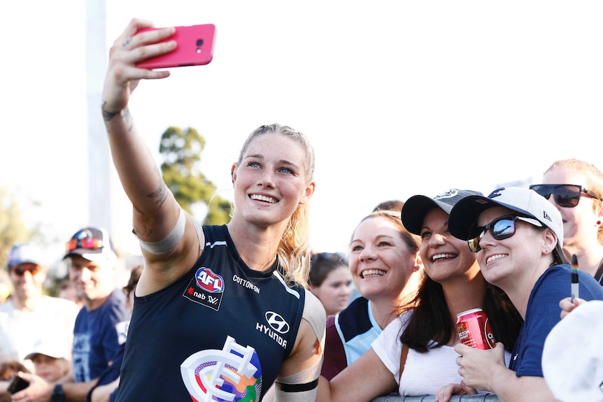 Tayla Harris sourit et lève un téléphone pour prendre un selfie avec des fans en marge