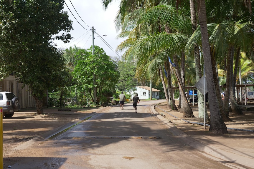 Two teenagers scoot down the middle of a road lines with palm trees and houses.