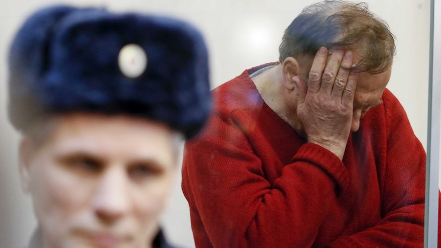 Sokolov in a red sweater has his hands buried in his face in despair, in front of him is a russian guard