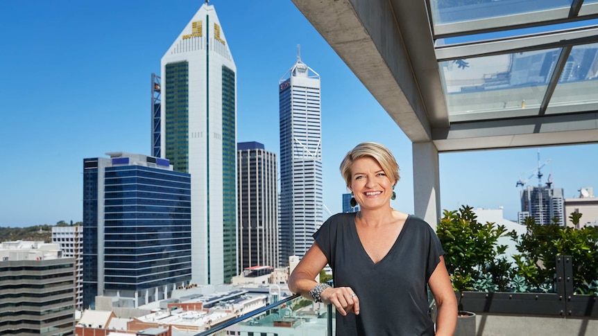 A woman with short blond hair, wearing a dark t-shirt, standing on a balcony overlooking the CBD.