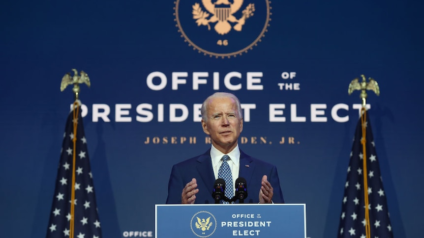 Joe Biden stands speaking at a podium in front of two American flags.