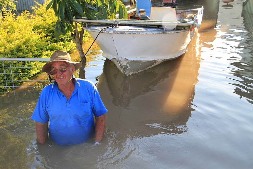 A man stands in waist-deep flood waters with a tinny boat behind him.