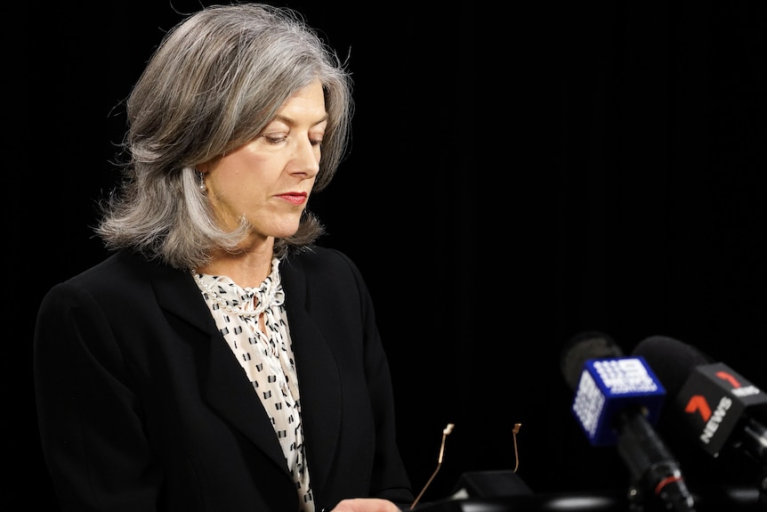 A woman with grey hair looks down at her glasses and microphones