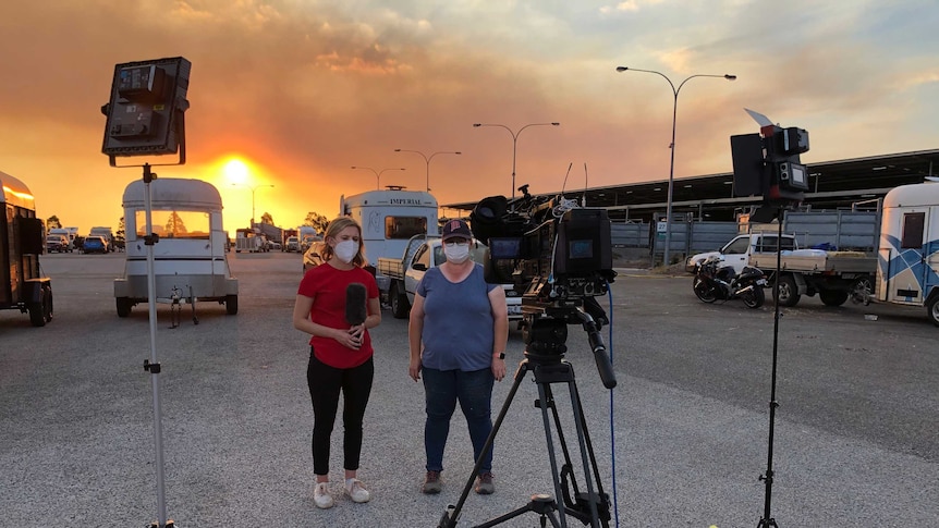 Journalist and woman wearing masks being filmed with horse floats and bright orange sky in the background.