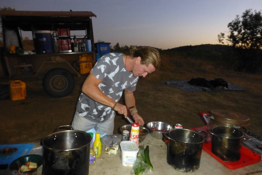 A man leans over a fold-out table with cooking ingredients, in the background a four-wheel drive.