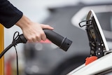 A hand holds a plug towards the open charging port of an electric car.