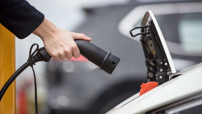 A hand holds a plug towards the open charging port of an electric car.