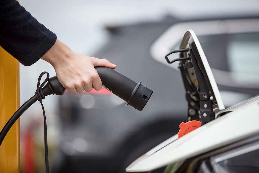 A hand holds a plug towards the open charging port of an electric car.