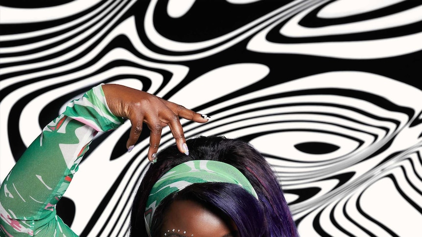 Yola wearing a green, white and red dress and matching headband striking a pose in front of a black and white patterned wall.