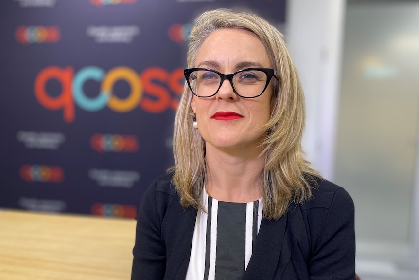 A blonde, bespectacled woman in formal clothes sits in a corporate office.