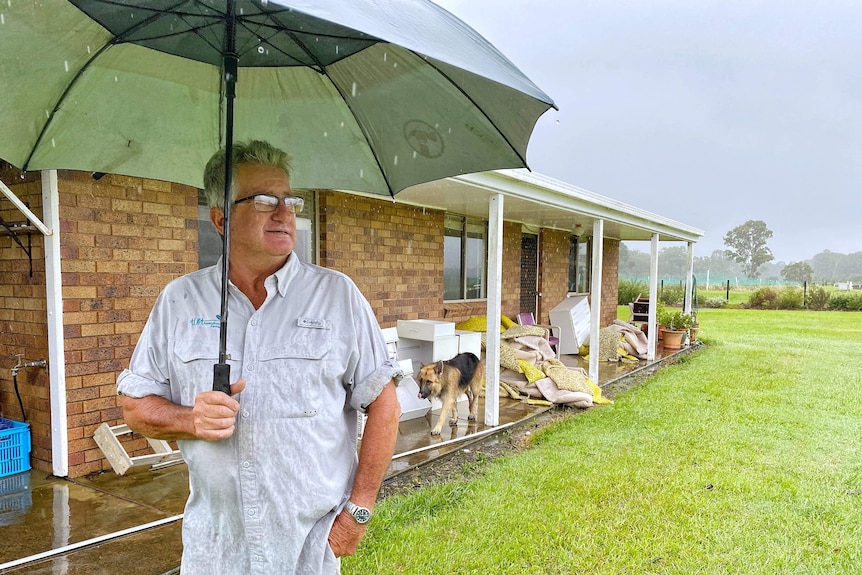 A man with an umbrella, stands in front of a house that has flood ruined furniture and carpet in front of it.