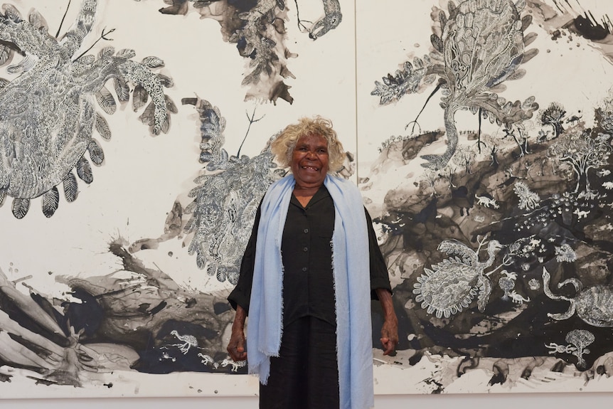 An First Nations woman in her 80s in black dress and blue shawl stands in front of an elaborate piece of art in black and white