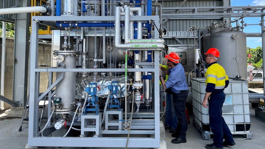 Two men wearing orange hard hats examine equipment and pipes of a biorefinery plant. 