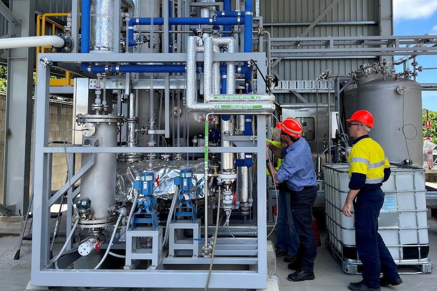 Two men wearing orange hard hats examine equipment and pipes of a biorefinery plant. 