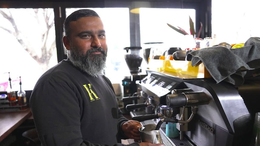A man with a thick beard steams milk in a metal jug at an espresso machine.