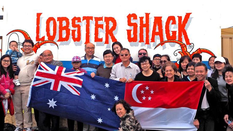 A group of people holding an Australian and a Singaporean flag in front of a wall reading Lobster Shack.