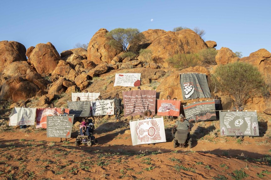 Paintings with words on them being held up by Aboriginal people on the side of a red hill