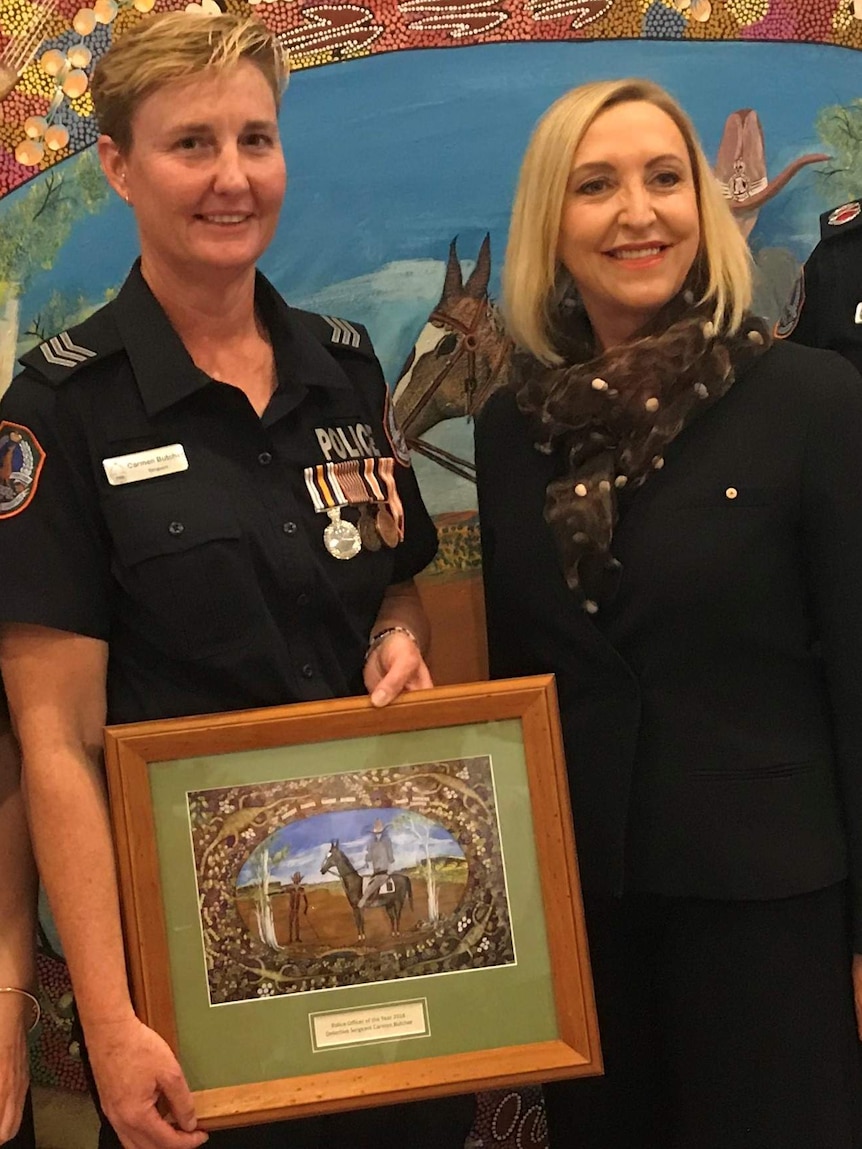 Police woman in blue uniform holding picture in frame standing next to woman with blond hair.
