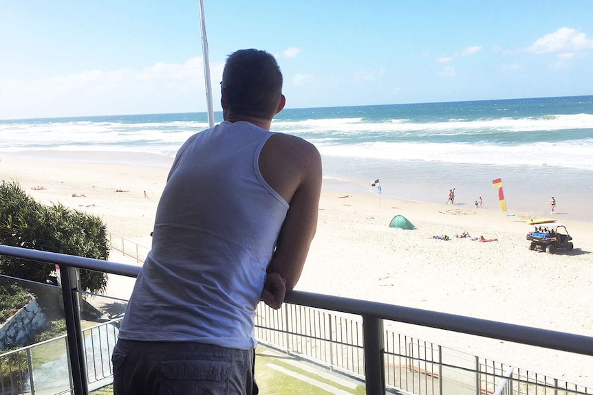 A man in a singlet looks down on a beach from a beachfront balcony