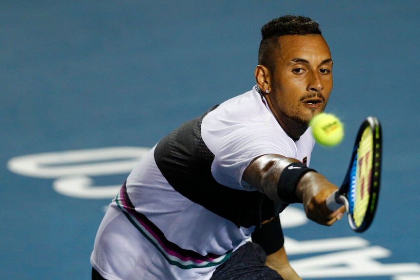 Nick Kyrgios wearing a black and white t-shirt reaching with his right hand for a tennis ball in the foreground
