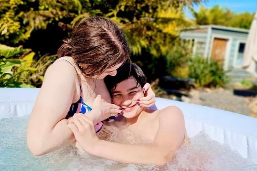 A girl cuddles a young boy in a pool. He's grinning at the camera.