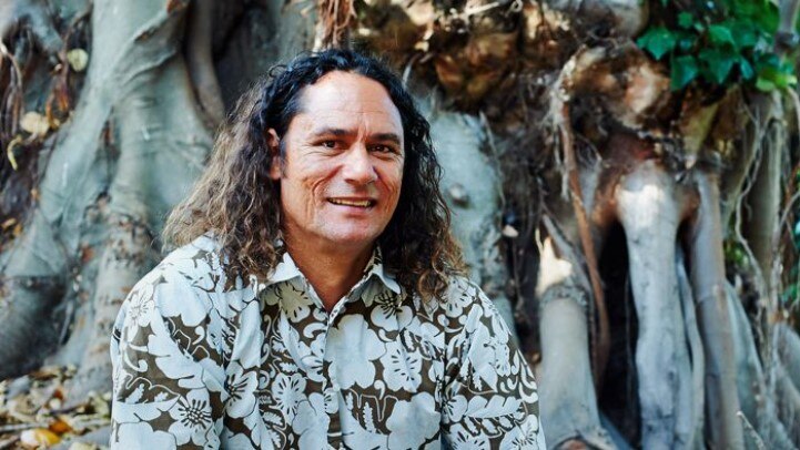 Man (Clarence Slockee) with long hair smiling at camera in patterned button up shirt. Sitting in front of large tree roots.