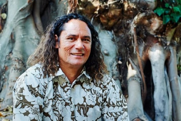 Man with long hair smiling at camera in patterned button up shirt. Sitting in front of large tree roots.