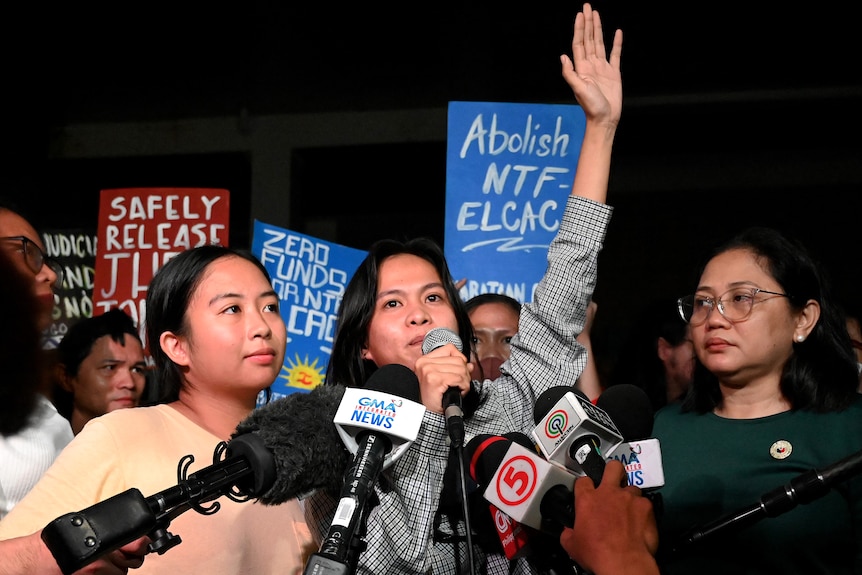 Two young women speak into microphones from news outlets while supporters hold up signs behind them.