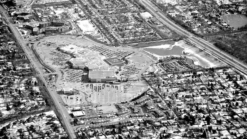 You see a black and white aerial view of a huge suburban shopping mall and carpark surrounded by low density housing.