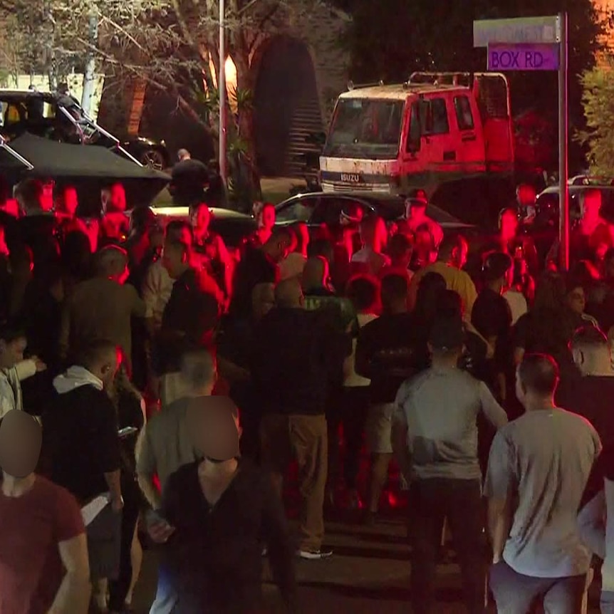 A crowd of people bathed in red light stand in a street at night.
