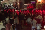A crowd of people bathed in red light stand in a street at night.